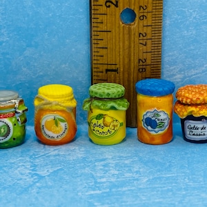 May include: Five miniature glass jars with colourful labels and lids. The jars are filled with different types of preserves, including orange marmalade, quince jelly, plum jam, and blackcurrant jelly. The jars are arranged in a row on a blue and white tiled surface.