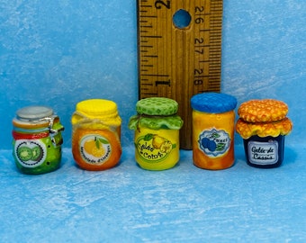 Può includere: Cinque vasetti di vetro in miniatura con etichette e coperchi colorati. I vasetti sono riempiti con diversi tipi di conserve, tra cui marmellata d'arancia, gelatina di cotogna, marmellata di prugne e gelatina di ribes nero. I vasetti sono disposti in fila su una superficie piastrellata blu e bianca.