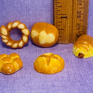 May include: Five miniature ceramic bread rolls in various shapes, including a ring, a round loaf, a braided loaf, and two smaller rolls. The rolls are painted in shades of brown and yellow, with some white details.