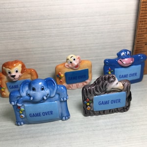 May include: Five ceramic figurines of different animals, each holding a sign that says "Game Over". The animals are a monkey, a zebra, an elephant, a hippopotamus, and a lion. The figurines are painted in bright colors and have a whimsical style.