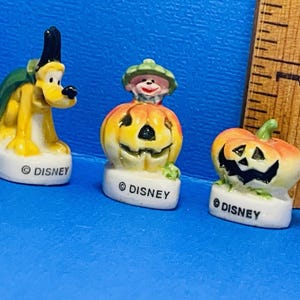 May include: Three small ceramic figurines of Disney characters dressed up for Halloween. A yellow dog wearing a green cape, a pumpkin with a face, and a pumpkin with a face and a green hat. Each figurine has a white base with the word "DISNEY" printed on it.