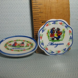 May include: Two small, white ceramic plates with blue and red trim. One plate has a blue and red floral design with a couple in traditional clothing and the name "Morgan". The other plate has a similar design with the name "Killian".