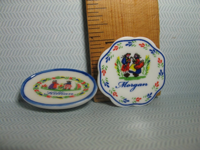 May include: Two small, white ceramic plates with blue and red trim. One plate has a blue and red floral design with a couple in traditional clothing and the name "Morgan". The other plate has a similar design with the name "Killian".