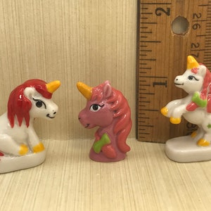 May include: Three small porcelain unicorn figurines. One is white with red and yellow accents, one is pink with green accents, and one is white with red, yellow, and green accents.