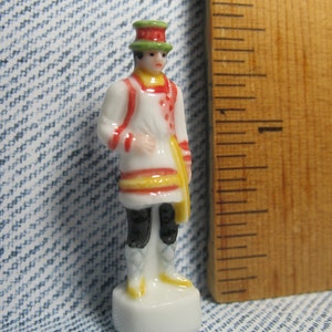 May include: A porcelain figurine of a person wearing a white, red, and yellow patterned shirt and a green and white hat. The figurine is standing on a white base.