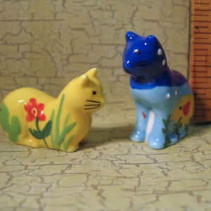 Puede incluir: Dos pequeñas figuras de gato de cerámica. Una es amarilla con una flor roja y hierba verde pintadas en el costado. La otra es azul con puntos blancos y un diseño de sol amarillo. Una regla de madera muestra una altura de 3,8 centímetros.