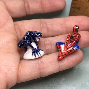 May include: Two small ceramic figurines of Spiderman and Venom. Spiderman is sitting with his legs crossed and wearing his signature red and blue suit. Venom is standing on a white base and is in a crouching position. Both figurines are painted in detail.