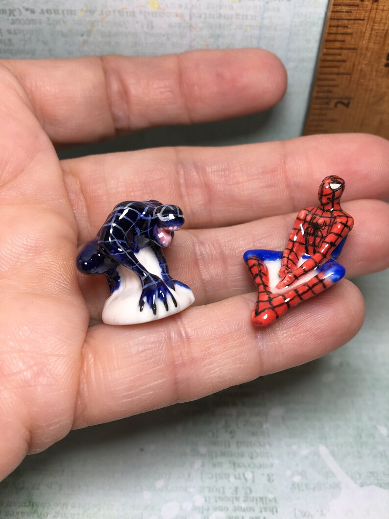 May include: Two small ceramic figurines of Spiderman and Venom. Spiderman is sitting with his legs crossed and wearing his signature red and blue suit. Venom is standing on a white base and is in a crouching position. Both figurines are painted in detail.