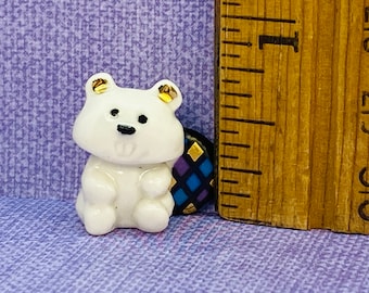 May include: A small white ceramic bear figurine with gold accents on the ears. The bear is sitting on a small, round, blue and gold patterned tile.