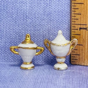 May include: Two miniature white and gold urns with lids and handles. The urns have a classic shape and are decorated with gold trim. The background is a light blue textured surface. A wooden ruler is next to the urns.