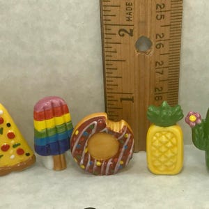 May include: A collection of seven small, colorful resin charms shaped like a red lobster, a pizza slice, a rainbow popsicle, a chocolate donut, a yellow pineapple, a green cactus with a pink flower, and a watermelon slice, next to a wooden ruler.