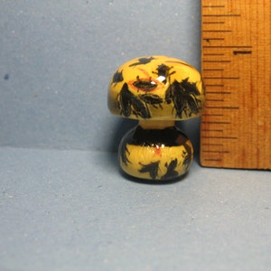 Pode incluir: Uma pequena figura de cogumelo de cerâmica com um design pintado em amarelo e preto. O cogumelo tem um chapéu arredondado e um caule curto.