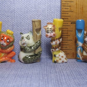 May include: Four ceramic animal whistle figurines. A lion, panda, giraffe, and monkey, each holding a whistle in their hand. The whistles are yellow, brown, blue, and yellow.
