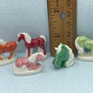 May include: Five miniature ceramic horse figurines. The horses are painted in various colors, including brown, red, pink, green, and white. The figurines are approximately 2 inches tall and are displayed on a blue and white patterned surface.