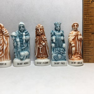 Peut inclure: Six petites figurines en céramique de saints, chacune avec un nom et un design différents. Les figurines sont peintes dans des tons de brun, bleu et blanc. Les figurines sont disposées en rangée sur une surface blanche.