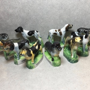 May include: A collection of ten miniature ceramic dog figurines, each depicting a different breed. The figurines are painted in realistic colours and feature intricate details. They are displayed on small green bases with the name of the breed written on each base.