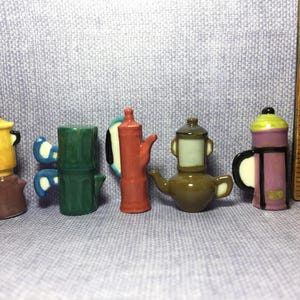May include: Five miniature ceramic coffee makers and teapots arranged in a row next to a wooden ruler. The items include a yellow and brown pot, a green cylindrical pot, a red pot, a brown teapot, and a purple and green French press.