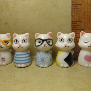 Puede incluir: Cinco figuritas de cerámica blanca de gatos con diferentes patrones y diseños. Cada gato tiene ojos negros y una nariz rosa. Los gatos llevan gafas, una camisa a rayas, un patrón floral, un patrón amarillo y negro y un corazón rosa.