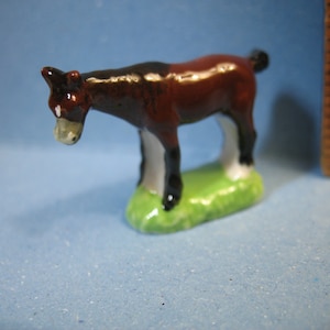 May include: A small brown and black ceramic figurine of a horse with white hooves standing on a green base.