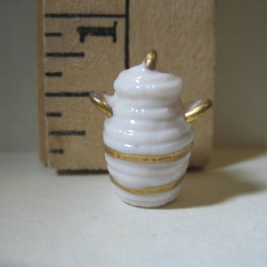 以下が含まれることがあります： 金色の縁取りと2つの取っ手が付いた小さな白い陶器の壺。壺は丸みを帯びた形をしていて、蓋が付いています。