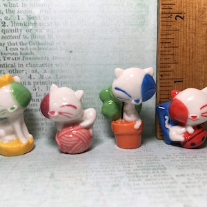 May include: Four ceramic cat figurines, each with a different color scheme and detail. One cat is holding a ball of yarn, another is holding a ladybug, another is in a pot with a green plant, and the last cat is wearing a crown.