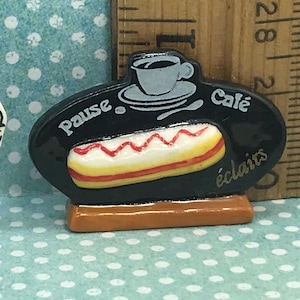Puede incluir: Un letrero ovalado negro con base marrón, que presenta un gráfico de una taza de café gris sobre un platillo encima de un éclair amarillo con glaseado rojo. El texto dice "Pause Café éclairs". Una regla muestra 3,8 cm de ancho.