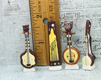 May include: Four miniature ceramic musical instrument figurines, including three round-bodied string instruments and one rectangular zither-style instrument, arranged next to a wooden ruler showing a height of approximately 2.5 inches.
