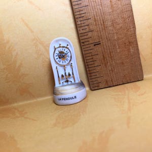 May include: White porcelain figurine of a grandfather clock with gold accents. The clock face has black Roman numerals and hands. The base of the clock reads "LA PENDULE".
