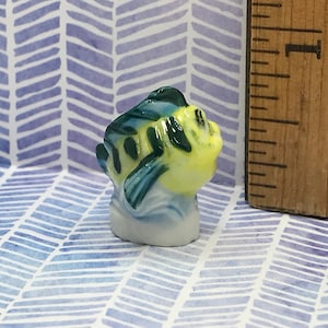 May include: A small ceramic figurine of a yellow and blue fish with green and blue markings. The figurine is about 1 inch tall and is sitting on a white background.