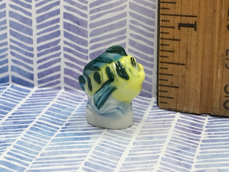 May include: A small ceramic figurine of a yellow and blue fish with green and blue markings. The figurine is about 1 inch tall and is sitting on a white background.