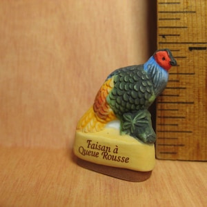 Peut inclure: Une petite figurine en céramique d'un faisan avec un motif peint vert, bleu et jaune. La figurine est posée sur un socle avec le texte "Faisan à Queue Rousse".