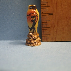 Puede incluir: Un pequeño jarrón de cerámica con un esmalte amarillo y marrón. El jarrón tiene un diseño floral y una figura estilizada en la parte delantera. El jarrón está sobre una superficie de madera.