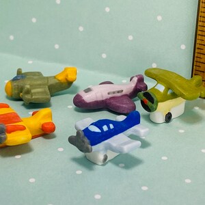 Könnte beinhalten: Fünf Miniatur-Spielzeugflugzeuge in verschiedenen Farben, darunter Gelb, Rot, Grün, Blau und Lila. Die Flugzeuge sind aus Keramik und haben bemalte Details.