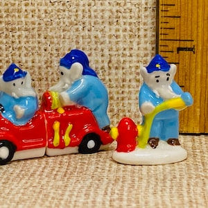 May include: Three ceramic figurines of elephants dressed as firefighters. One elephant is driving a red fire truck, another is holding a hose, and the third is standing next to the truck.