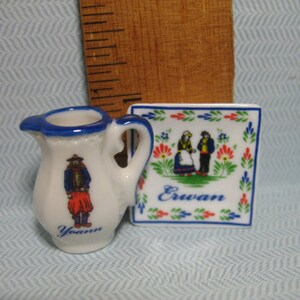 May include: A white ceramic pitcher with a blue rim and a blue and red illustration of a person in traditional clothing. The pitcher is next to a white ceramic square tile with a blue and red border and a blue and red illustration of two people in traditional clothing. The tile has the word "Erwan" written in blue.