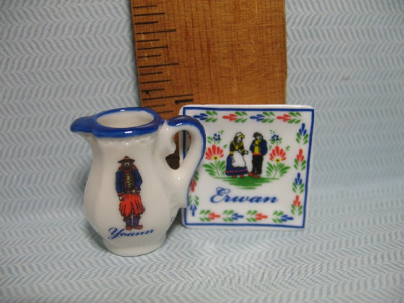 May include: A white ceramic pitcher with a blue rim and a blue and red illustration of a person in traditional clothing. The pitcher is next to a white ceramic square tile with a blue and red border and a blue and red illustration of two people in traditional clothing. The tile has the word "Erwan" written in blue.