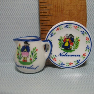 May include: A white ceramic creamer and saucer set with blue and red trim. The creamer features a painted image of a woman in a blue dress and a pink skirt. The saucer has a painted image of two people in traditional clothing and the word "Nolwenn".
