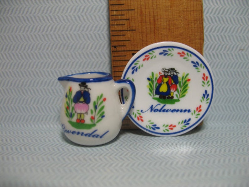 May include: A white ceramic creamer and saucer set with blue and red trim. The creamer features a painted image of a woman in a blue dress and a pink skirt. The saucer has a painted image of two people in traditional clothing and the word "Nolwenn".
