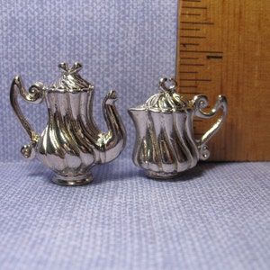 May include: Two silver-toned metal teapots with a decorative swirl design. The teapots are small and appear to be charms or ornaments.