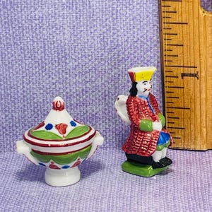 Pode incluir: Miniaturas de cerâmica de um pote com tampa e um homem de casaco vermelho. O pote é branco com detalhes verdes, vermelhos e azuis. A figura masculina usa um casaco vermelho, calças azuis e um chapéu amarelo. Uma régua de madeira está ao fundo.