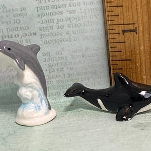 May include: Two ceramic dolphin figurines. One is a grey dolphin leaping out of the water, the other is a black and white orca dolphin.