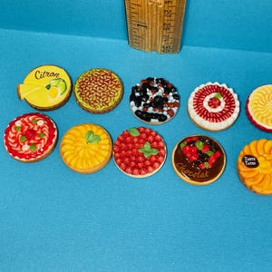 Może przedstawiać: Kolekcja miniaturowych, okrągłych, dekoracyjnych tart z różnymi dodatkami, w tym owocami i czekoladą. Jedna tarta ma napis "Citron", a druga "Tarte Tatin". Tarty znajdują się na niebieskiej powierzchni.