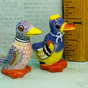 Könnte beinhalten: Zwei kleine Keramik-Entenfiguren. Eine Ente ist blau, gelb und schwarz mit einem weißen Band um den Hals. Die andere Ente ist mehrfarbig mit blau, gelb, rot und weiß. Beide Enten haben einen roten Schnabel und orangefarbene Füße.