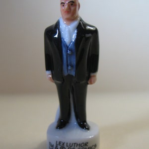 K&ouml;nnte beinhalten: Eine Porzellanfigur von Lex Luthor, einem kahlk&ouml;pfigen Mann im schwarzen Anzug und blauer Weste. Die Figur steht auf einem wei&szlig;en Sockel mit dem aufgedruckten Text "TM LEX LUTHOR (C) DC COMICS".
