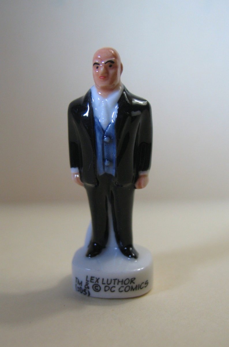 K&ouml;nnte beinhalten: Eine Porzellanfigur von Lex Luthor, einem kahlk&ouml;pfigen Mann im schwarzen Anzug und blauer Weste. Die Figur steht auf einem wei&szlig;en Sockel mit dem aufgedruckten Text "TM LEX LUTHOR (C) DC COMICS".