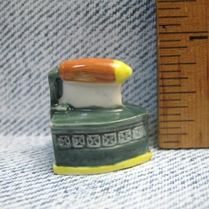 May include: A small ceramic figurine of a vintage iron with an orange and yellow handle, a white body, and a green base with a decorative pattern.