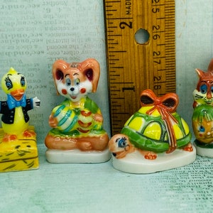 May include: Four small ceramic figurines, each depicting a different animal. A yellow duck with a blue bow tie, a brown rabbit holding two Easter eggs, a green turtle with a red bow, and a brown squirrel holding an Easter egg.