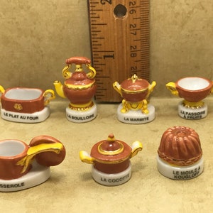 Może przedstawiać: Zestaw miniaturowych ceramicznych figurek przedstawiających różne przybory kuchenne. Figurki są pomalowane w odcieniach brązu i żółci i mają francuskie nazwy dla każdego przedmiotu, takie jak "La Bouilloire" i "Le Plat Au Four".