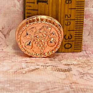 May include: A rose gold-toned metal button with a raised design of grapes and leaves.