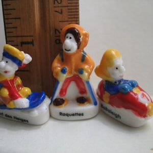 May include: Three ceramic figurines depicting winter sports. The first figurine is a person wearing a blue and yellow hat and red and yellow clothing, riding a blue and white sled. The second figurine is a person wearing a red and orange jacket and blue pants, holding two sticks. The third figurine is a person wearing a blue and yellow hat and blue and red clothing, riding a red and yellow bobsled. The figurines are labelled "Surf des Neiges", "Raquettes", and "Bobsleigh".